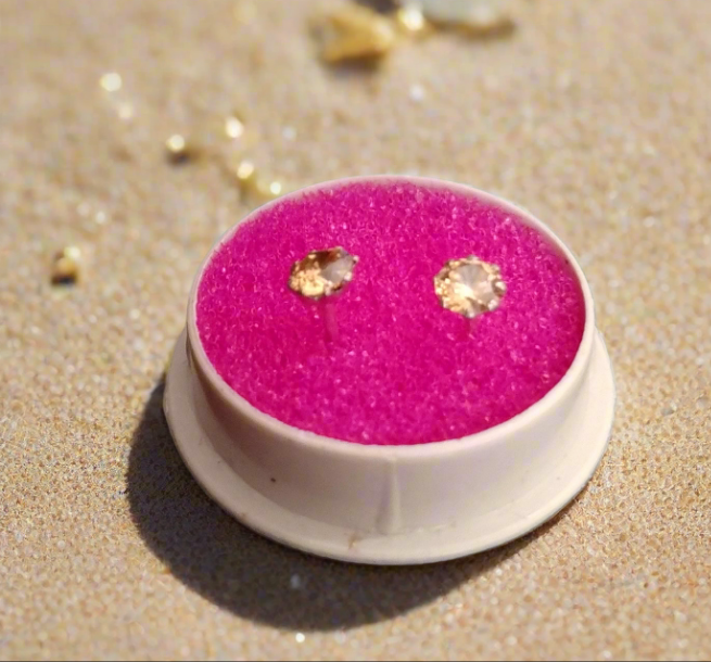 Gold earrings on a pink cushion in a white holder on sand