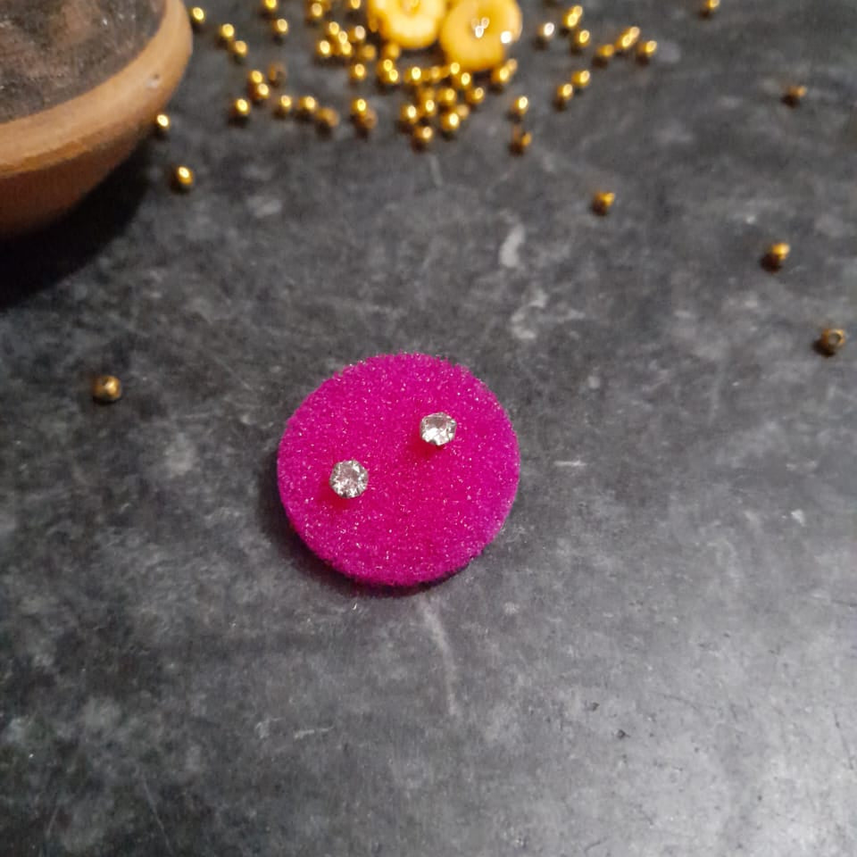 Pink earring with a clear gemstone on a dark surface with small yellow objects and a wooden item.